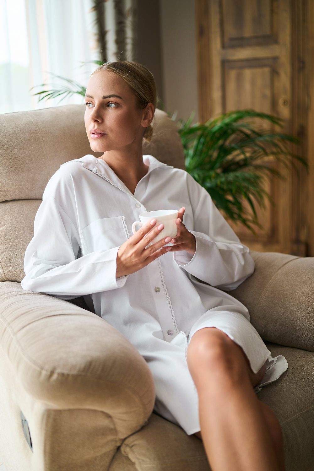 Kobieta w białej koszuli nocnej siedząca na kanapie, trzymająca filiżankę w dłoniach