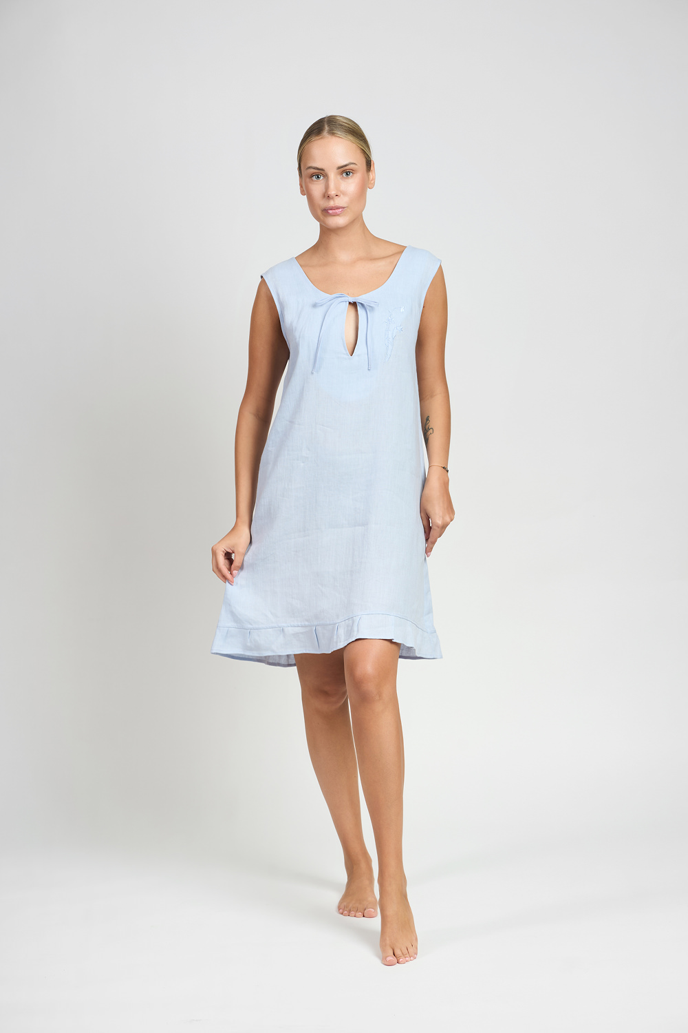 Kobieta w błękitnej lnianej koszuli nocnej z wiązaniem przy dekolcie idąca na jasnym tle