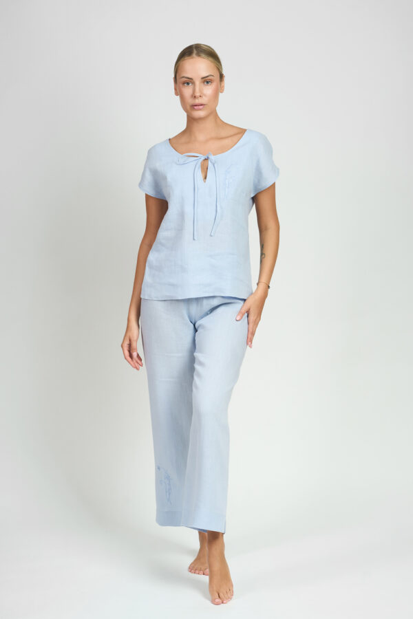 Kobieta w pełnym błękitnym lnianym komplecie z bluzką z wiązaniem przy dekolcie na jasnym tle