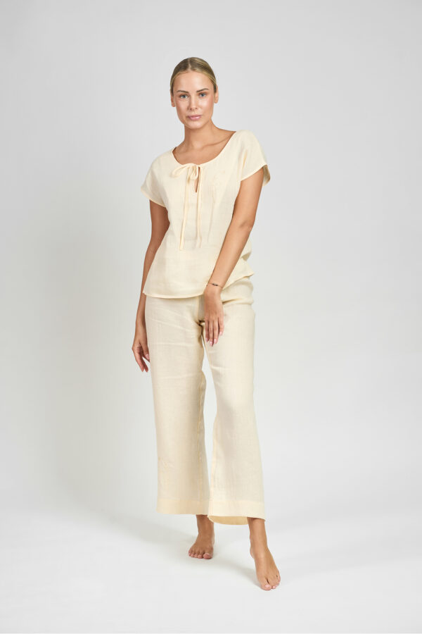 Kobieta w kremowym lnianym komplecie z bluzką z wiązaniem przy dekolcie, stojąca na jasnym tle