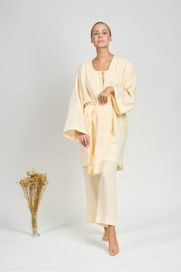 Kobieta w kremowym lnianym szlafroku i spodniach, obok wazon z suszonymi kwiatami