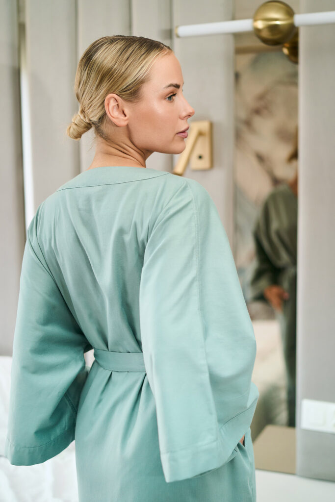 Blond kobieta w szałwiowym szlafroku stojąca profilem przed lustrem, z włosami upiętymi w kok